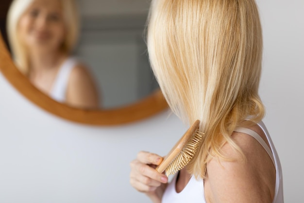 Rear view of middle aged woman brushing hair with wooden hairbrush looking at mirror in bathroom at