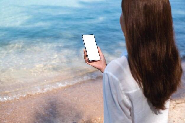 Rear view mockup image of a woman holding mobile phone with\
blank desktop screen while sitting on the beach