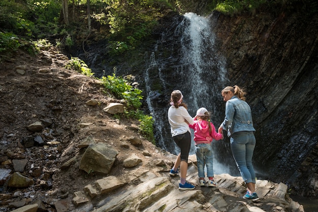 Rear view mother and two little daughters are standing on mountain and enjoying bewitching view of waterfall while trekking through mountains on sunny summer day. Concept of tourism with children