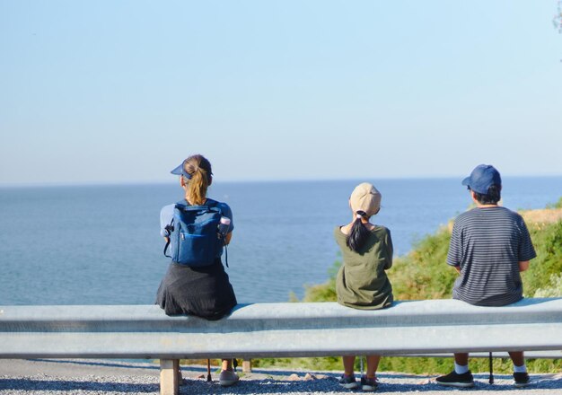 Rear view of mother with daughter and son sitting on railing while looking at sea against sky