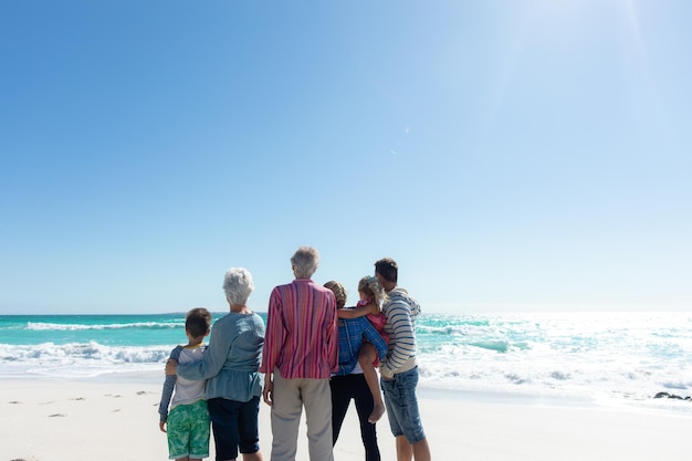 Rear view of a multi-generation Caucasian family standing on the beach with blue sky and sea in the background, embracing and looking away