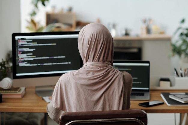 Rear view of Muslim female programmer in hijab sitting in front of computer screen