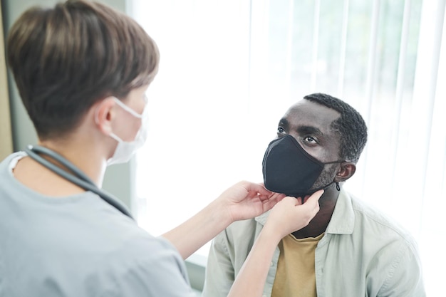 Вид сзади медсестры в маске, касающейся шеи афроамериканского пациента во время осмотра его здоровья в клинике