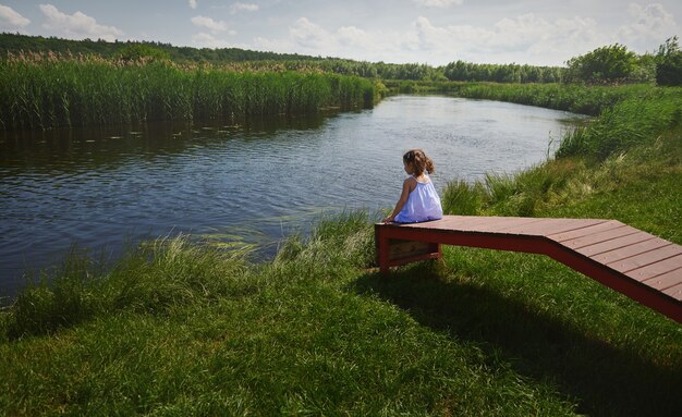 Фото Вид сзади милая девочка с двумя хвостиками, сидящая на деревянном пирсе на берегу реки и смотрящая на противоположный берег ребенок проводит время на природе на свежем воздухе в сельской местности