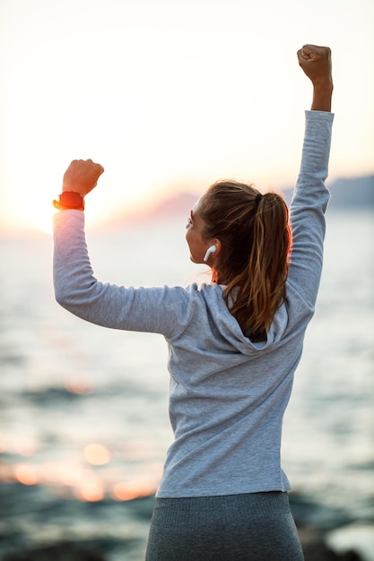 写真 幸せな若い女性の背面図は、海のビーチの近くでトレーニングの準備をしていて、夏の夕日や日の出を楽しんでいます。