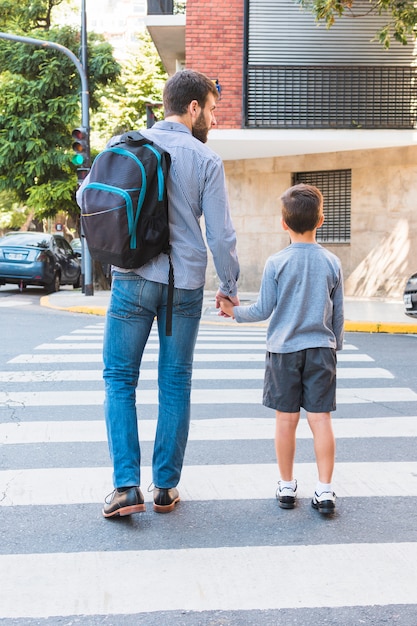 写真 彼の息子と横断歩道を歩いてスクールバッグを運ぶ男の背面図