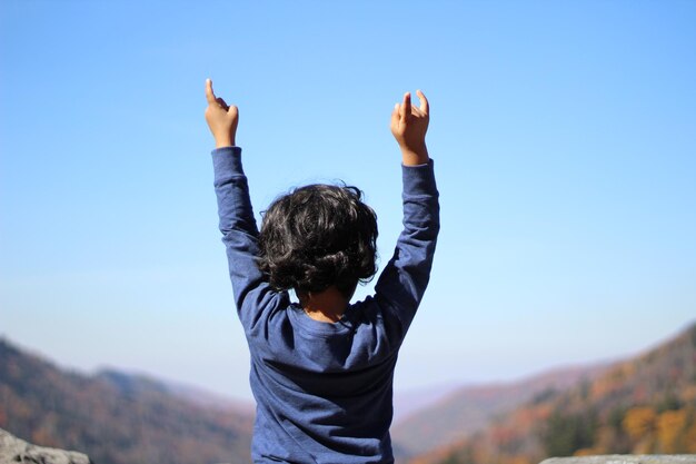 写真 明るい空に背を向けて腕を上げた少年の後ろの景色