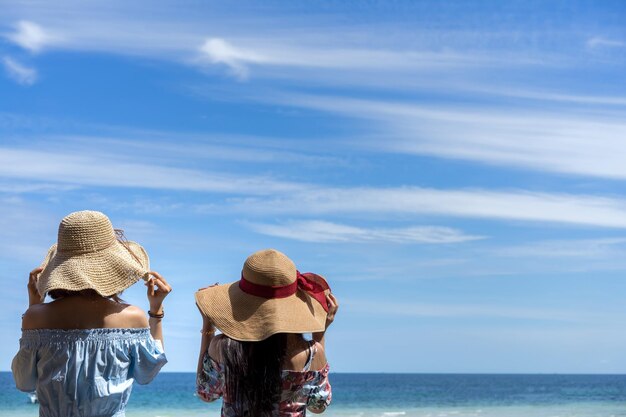 写真 晴れた日に青い空に照らされて海辺に立っている帽子をかぶった友達の後ろの景色