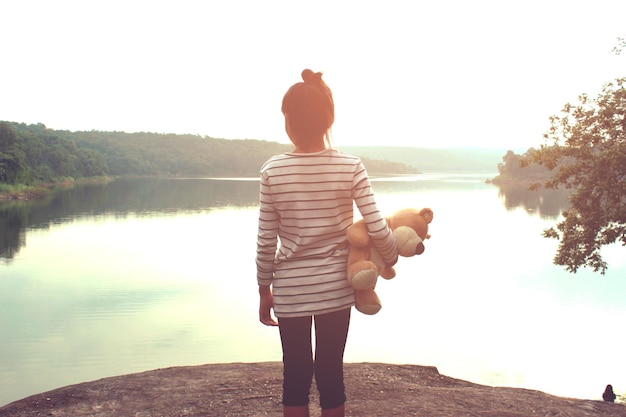 写真 空の向こうの湖の岩の上に立っているテディベアの女の子の後ろの景色