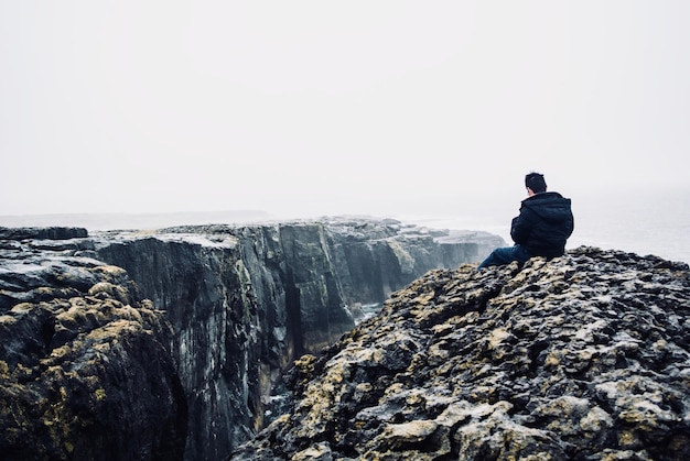 Фото Задний вид человека, сидящего на скале на фоне неба