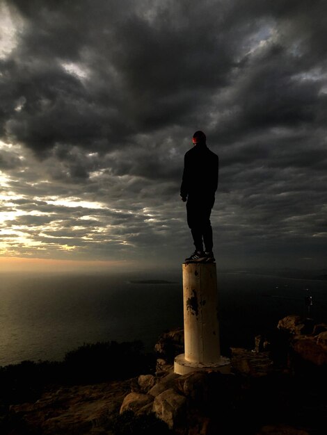 사진 해가 지는 동안 하늘 을 배경 으로 바위 위 에 서 있는 사람 의 뒷면