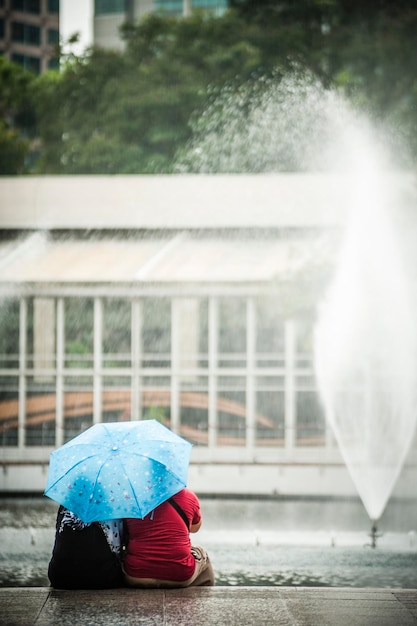 写真 街の噴水のそばで傘をかぶって座っている人々の後ろの景色