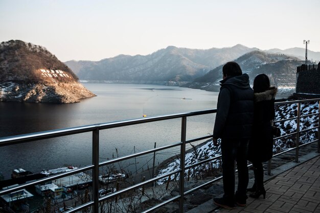 写真 空の向こうの山で湖を越えた橋のレールに立っている人々の後ろの景色