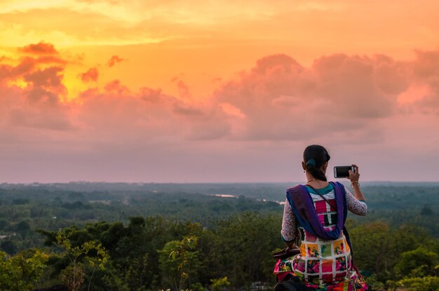 写真 夕暮れの空を背景に携帯電話で写真を撮っている女性の後ろの景色