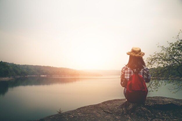 写真 夕暮れの湖を見ながら岩の上に座っている女性の後ろの景色