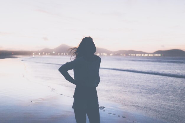 写真 夕暮れの際にビーチに立っている女性の後ろの景色