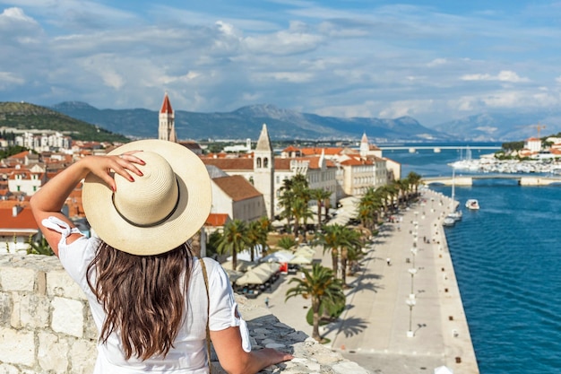 写真 トロギル (クロアチア) の美しい海辺の町を眺めている城壁に立っている女性の後ろの景色
