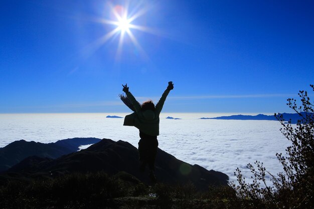 写真 青い空を背景に山の上に立っている腕を上げた女性の後ろの景色