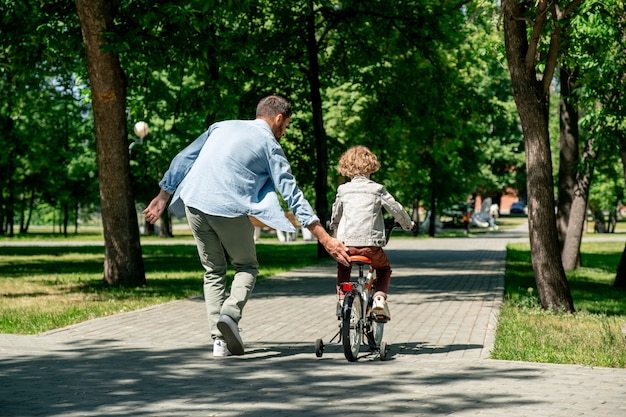 緑の芝生の間の公共公園の道路に沿って移動しながら、彼のかわいい幼い息子が自転車に乗るのを手伝っている若いアクティブな父親の背面図