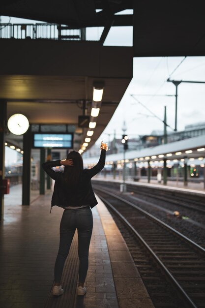 写真 鉄道駅のプラットフォームに立っているストリングライトを握っている若い女性の後ろの景色