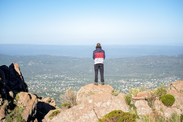 Rear view of one man standing on a rock in the edge of mountain\
overlooking a town against skyline