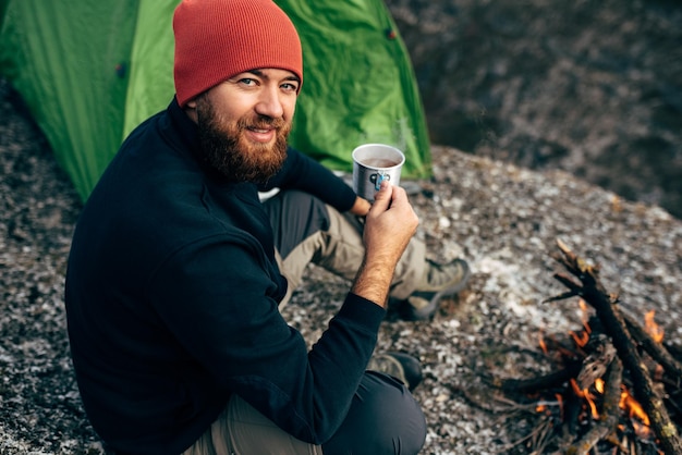 Rear view outdoor shot of young breaded male drinking hot beverage in mountains Traveler man in red hat sitting near to camping tent and bonfire holding in hands a mug of tea after hiking Travel