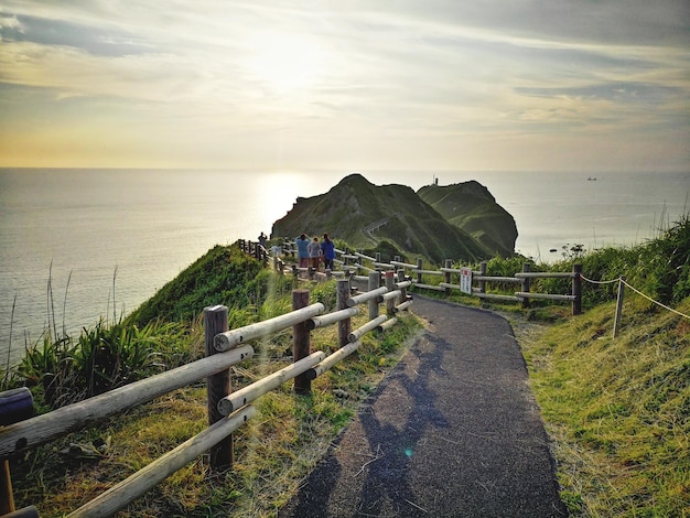 夕暮れの空に照らされて海に沿って歩道を歩く人々の後ろの景色