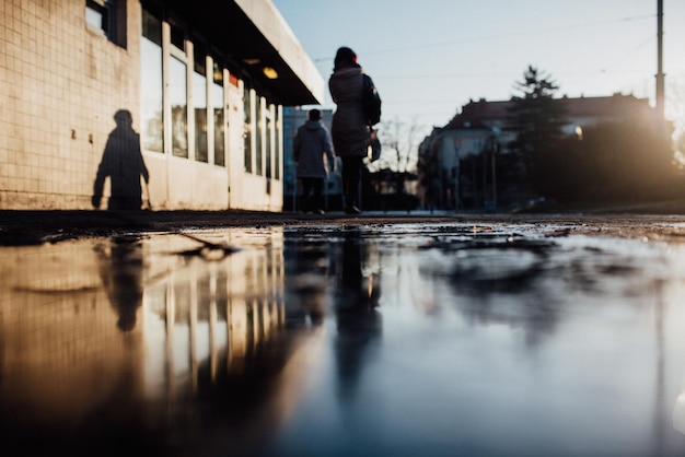 rear view of people walking on puddle on footpath in city against sky