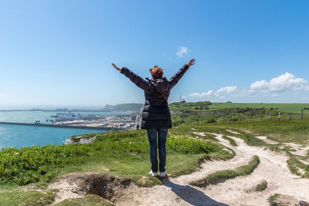 Rear view of person with arms outstretched standing on hill by sea against sky