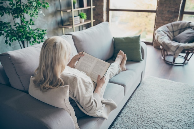 Rear behind view photo of blond aged granny lying comfy sofa divan reading favorite historic novel book nice free time wear beige pastel clothes flat living room indoors