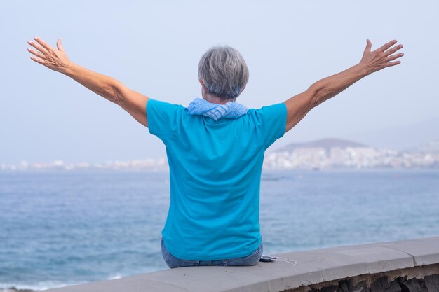 Rear view portrait of happy senior woman wearing blue tshirt and scarf enjoying outdoors freedom and vacation at sea Nice female looking at horizon with outstretched arms