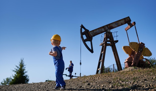 Rear view of preschool guy in protective helmet standing on stone road and pointing finger up to oil pump jack Petroleum borehole adult mechanic and blue sky on the blurred backdrop