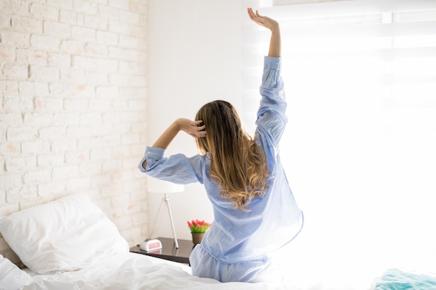 Rear view of a pretty young woman sitting on her bed and stretching her arms after waking up