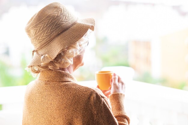 屋外でコーヒーカップと休憩を楽しんで帽子とサングラスと黄色のリラックスした巻き毛の年配の女性の背面図