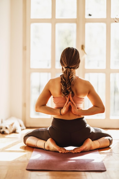 Rear view of a relaxed young woman practicing yoga in Back Namaste pose at home.