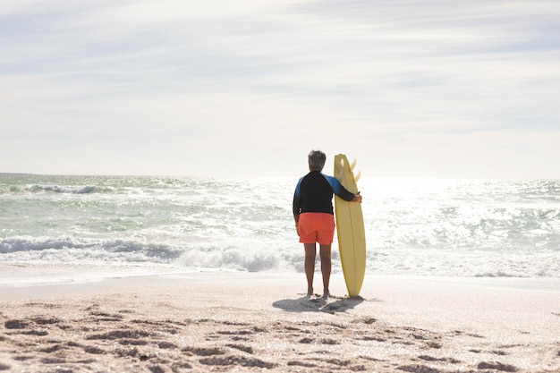 Rear view of senior biracial woman standing with surfboard on shore at beach during sunny day