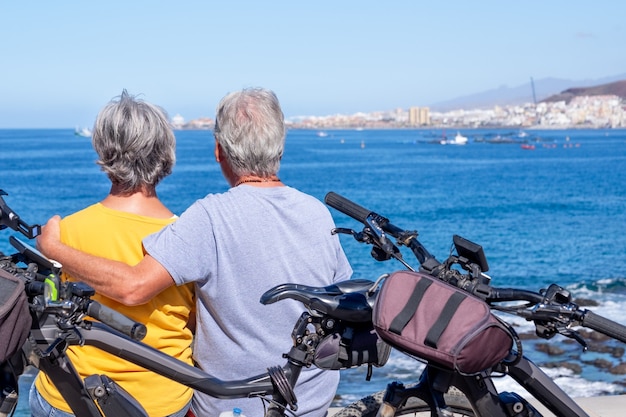 Vista posteriore della coppia senior seduta sulla scogliera in escursione in mare con le loro biciclette, guardando l'orizzonte. pensionati attivi che godono di uno stile di vita sano