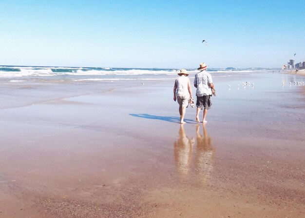 Rear view of senior couple walking at beach against blue sky