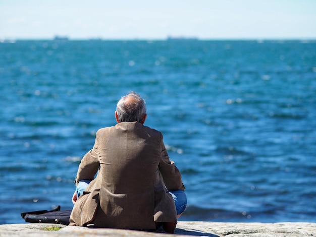 Rear view of senior man sitting on rock while looking at sea