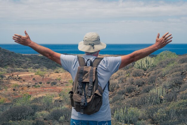 Rear view of senior man traveler enjoying outdoors trekking, looking at horizon over sea with open arms