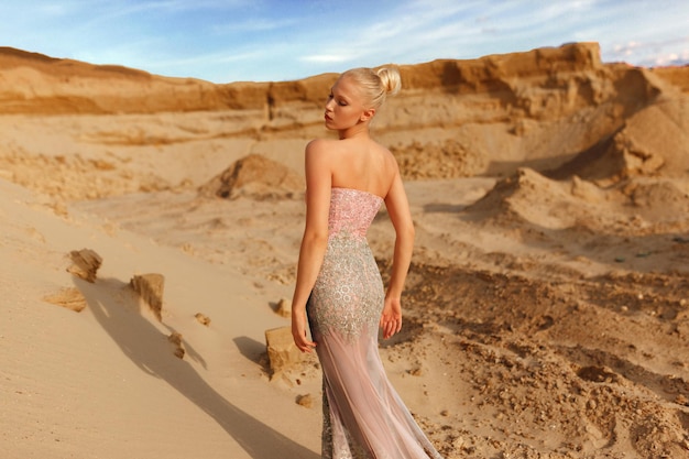 Rear view of a sensual young blonde woman in the desert in maxi gold dress posing in desert, at sunset background.