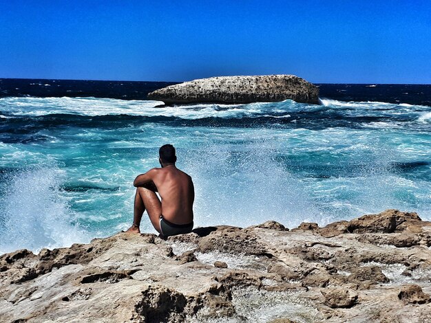 Rear view of shirtless man on rock at beach against sky