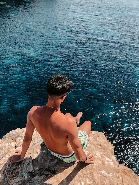 Rear view of shirtless man sitting on rock by sea