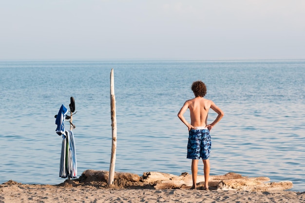 Photo rear view of shirtless teenage boy with hand on hip standing at beach against sky