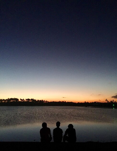 Photo rear view of silhouette friends sitting at lakeshore against sky during sunset