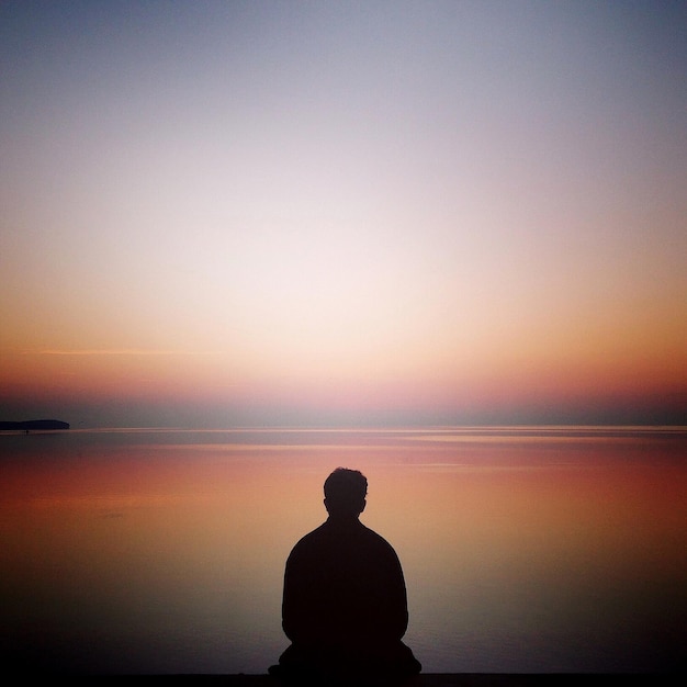 Foto vista posteriore di un uomo a silhouette seduto sulla spiaggia contro un cielo limpido