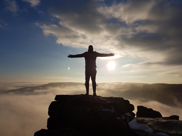 Foto vista posteriore di un uomo a silhouette con le braccia stese in piedi su una roccia contro il cielo durante il tramonto