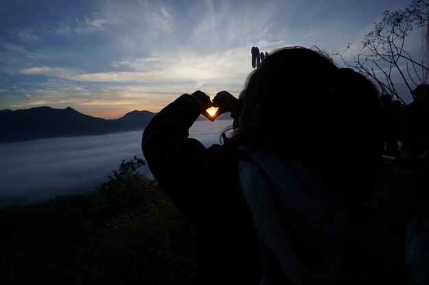 夕暮れの空に反して山の上で心の形をしているシルエットの人物の後ろの景色