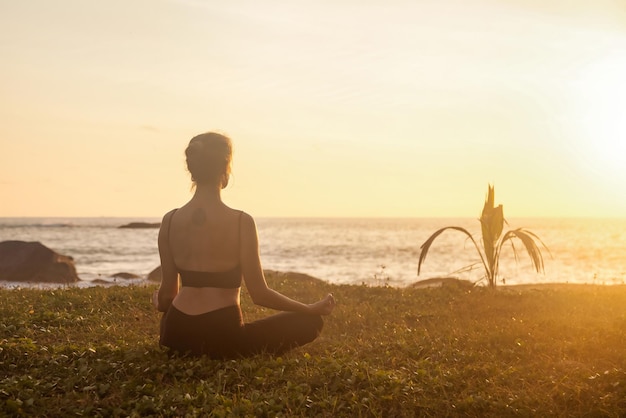 La donna esile della siluetta di retrovisione fa yoga sulla costa del mare tropicale o sulla spiaggia dell'oceano all'aperto sul tramonto