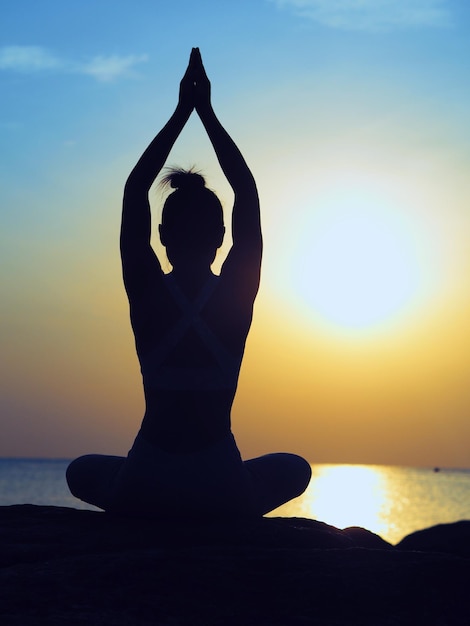 Rear view of silhouette woman meditating while sitting by sea against sky during sunset